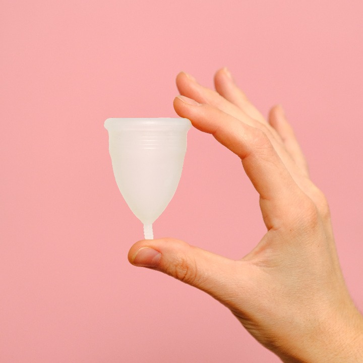 a hand holding a clear menstruation cup against a blush pink background