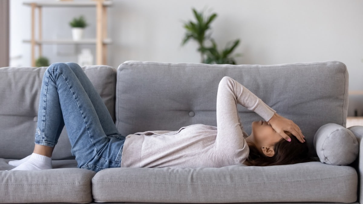 A stock image of a young woman lying on a couch with her hands on her face, looking frustrated