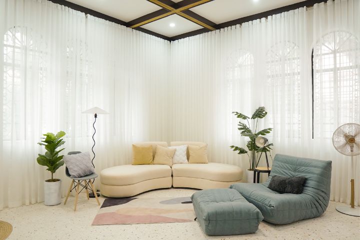 A living room featuring a comfy couch, rug, and bean bag chair combo, plus several tall windows and sheer curtains to let in tons of natural light.