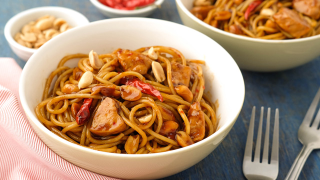 kung pao chicken pasta in a white bowl with peanuts and chili in condiment dishes