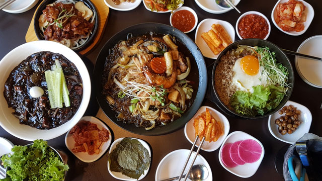 flat lay of different korean food including jjajangmyun, bibimbap, jeyuk bokkeum, japchae, and various banchan