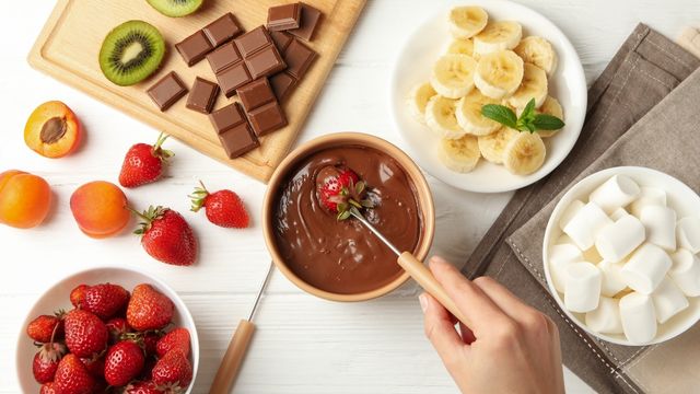 strawberring getting dipped in melted chocolate, surrounded by blowls of strawberry, bananas, and marshmallows